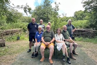 A photo of eight people sitting on or standing around a circular bench in the middle of a wildlife garden. They are smiling at the camera.