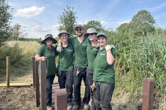 Five conservation trainees in matching green shirts and sun hats smiling with arms around each other while standing on a newly installed bridge amid reeds and grasses