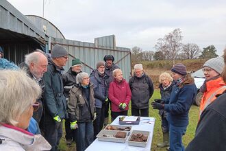 A group in winter coats and hats gathered outdoors around a table with trays of pottery and metal finds, listening to a woman in a beanie who gestures as she explains during an archaeology dig.