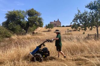 A volunteer wearing a wide-brim hat and gloves operating a mower to trim tall dry grass in a sunlit meadow, with a brick farmhouse on a hillside and scattered trees in the background.