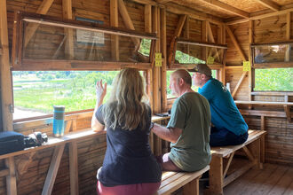 Three people in a birdwatching hide talking and looking out of the window