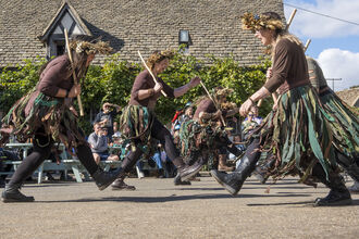 A photo showing a group of Morris dancers performing in an outdoor square with a building behind and people sat around watching on picnic benches