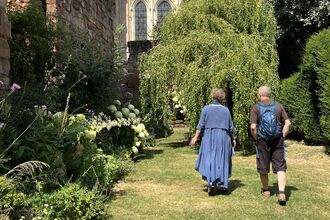 Two people walking away along a sunlit grassy path in a lush walled garden, passing hydrangeas and tall perennials toward a willow tree and stained-glass church windows set in old stone walls.