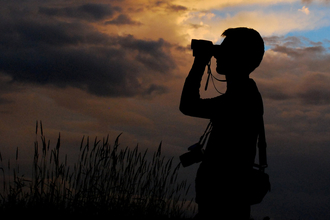 Silhouette of a person looking through binoculars against a sky. The sun is setting and the sky, with a hint of blue, is lit with a warm golden glow.