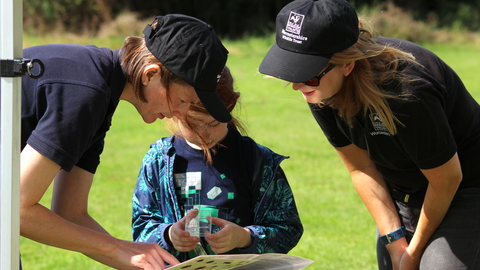 2 young women with WWT logos helping a young girl to identify an insect by Lauren Roberts
