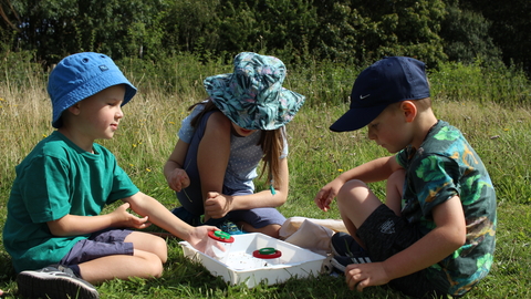 Three children with minibeast pots in a grassy field by Lauren Roberts