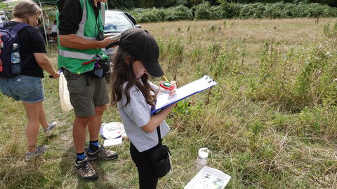 A photo of a girl standing in a meadow holding a clipboard doing bug hunting with two adults standing behind