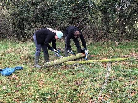 Two people handling a fence post on the ground with fencing leading to the foreground