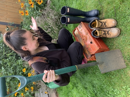 Woman with dark her and a rucksack on her back is looking down and wondering how to carry a pair of boots, a pair of wellies, a spade and an orange tool box