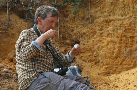 Man sitting in front of a small sandy cliff looking at an insect in a tube by Wendy Carter