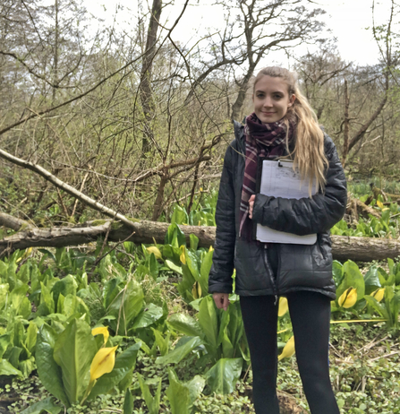 Woman with blond hair standing amongst skunk cabbage, an invasive species, holding a clipboard