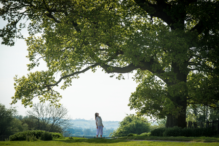 Woman standing underneath and looking up into the canopy of a tree with a wide landscape behind them both by Matthew Roberts