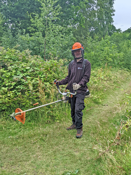 Man in protective workwear with a brushcutter on the side of a woodland path, smiling and looking at the camera
