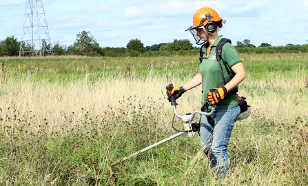 Woman in a field on a sunny day using a brushcutter to cut long grass by Lauren Roberts