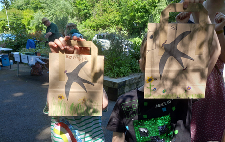 Two children holding up paper bags that they've decorated with swifts and wildflowers by Liz Yorke
