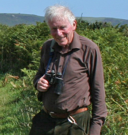 Man with white hair smiling at the camera as he stands with a pair of binoculars surrounded by nature