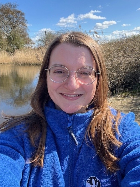 Woman wearing glasses and long brown hair smiling at the camera - it's a sunny day and there is a pond or lake behind her