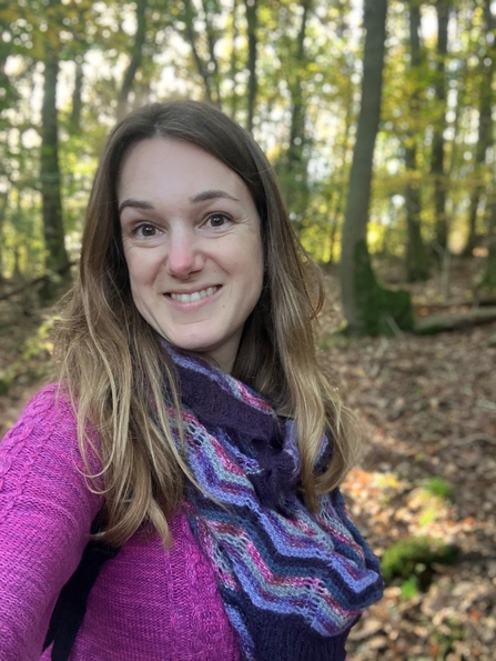 Woman with long brown hair smiling at the camera, she's in a sunlit woodland