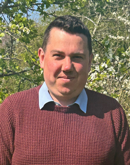 A man with dark hair smiling at the camera on a sunny day