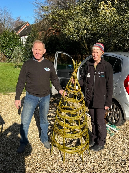 A man and a woman standing in the sunshine with a woven willow obelisk between them
