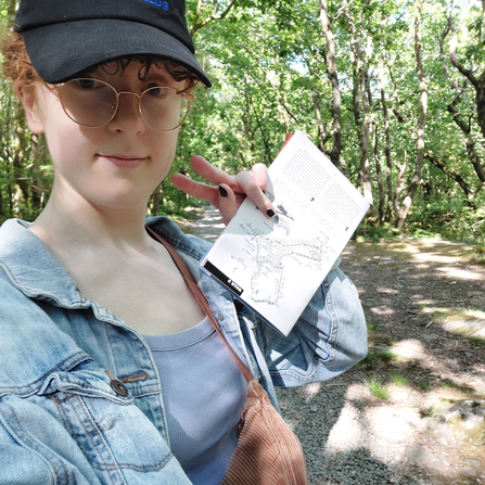 A woman with a peaked cap and glasses - she's standing in a woodland and is looking at the camera as she takes a selfie