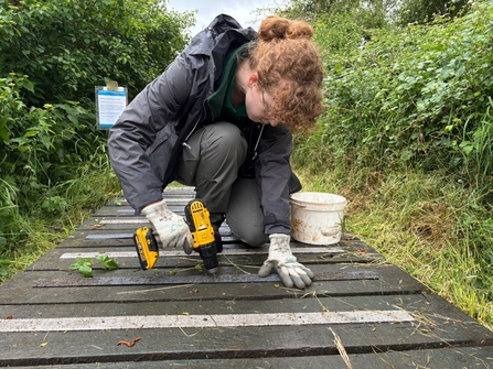 A person with long curly hair tied back is drilling anti-slip strips onto a boardwalk