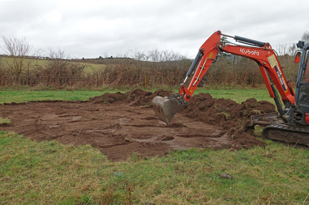 A digger scraping top soil from a field and building it up into a bank on the edge of the excavation; it's a winters day white a white sky but it doesn't look very cold