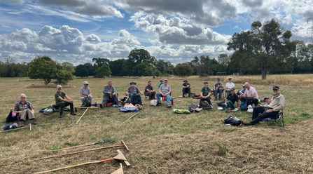 A group of people sitting in a semi-circle - they're having lunch after a morning raking mown grasses in a big meadow. It's a late summer day with blue sky and fluffy clouds.