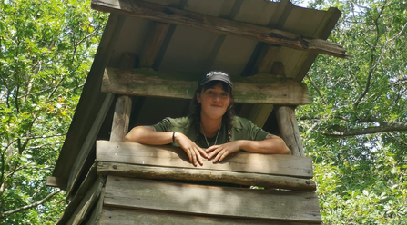 A smiling woman with a cap on is leaning out of the window of what looks like a treehouse - it's a sunny day and there's a tree canopy behind her