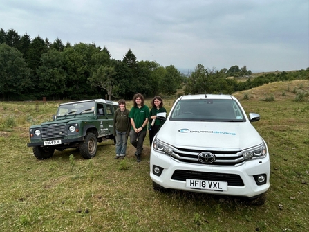 Three people standing between two off-roading vehicles; they're off-road with a woodland to the let and in a lumpy landscape