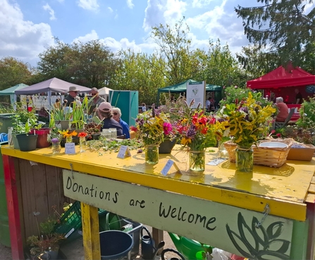 A 'donations are welcome' stall that's got lots of jars with cut flowers on it. There are other stalls in the background.