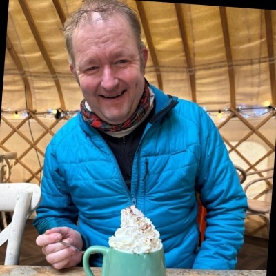 A man in a blue coat is laughing at the camera as he is about to tuck into a hot chocolate topped with whipped cream