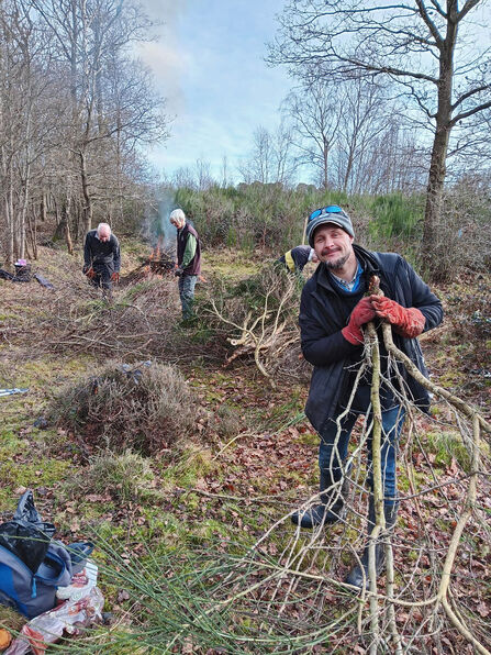 A number of volunteers are clearing scrub in the winter on a heathland. One is holding part of a tree and smiling at the camera.