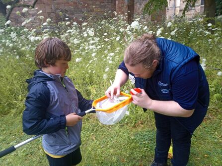 An adult with long hair tied back and a gilet is helping a child look into a net - they're also holding a bug pot