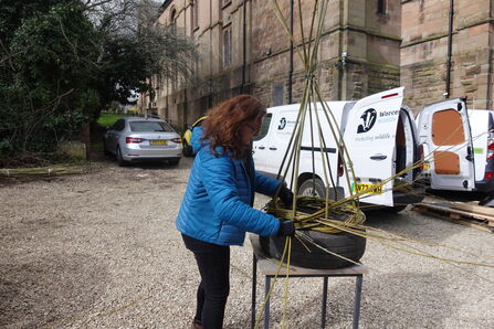 A person making a willow obelisk using an old tyre placed on a table with vehicles in the background