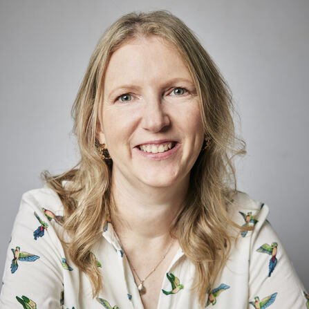 A person with long blonde hair smiling at the camera wearing a blouse with birds on