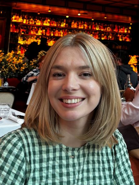 A head and shoulder photograph of a person with a blonde bob smiling and wearing a green gingham top