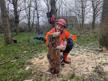 A man in high vis protective clothing and a helmet is crouching behind a newly felled tree stump and smiling at the camera