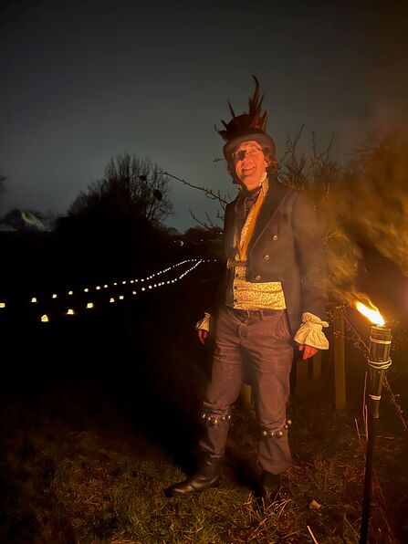 A man dressed as a Wassail Butler - with bells around knees and feathers in a top hat; it's dark and there's a torch alight in the foreground with trees in the background