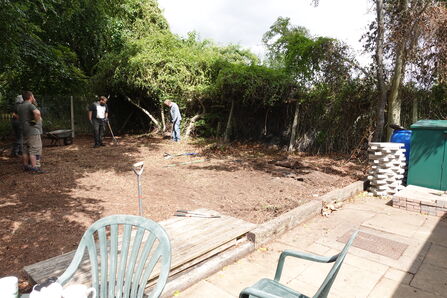 Four people holding spades working to clear an outdoor dirt space surrounded by a fence and trees