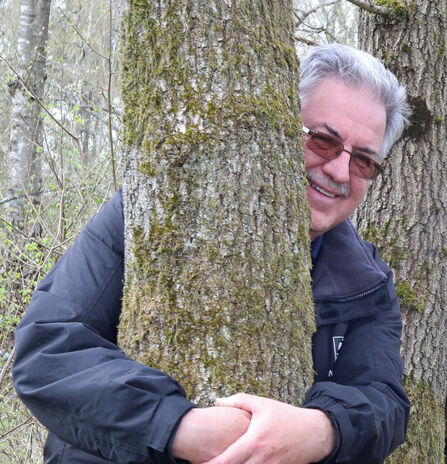 Bob Gillmor - a man with grey hair, sunglasses and a black jacket is hugging a tree and smiling at the camera