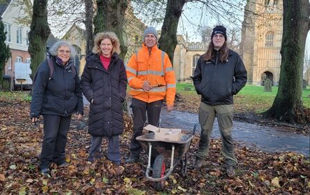 Four people standing by a wheelbarrow with trees and St Lawrence behind them