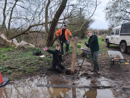 A photo of three trainees smiling at the camera standing and sitting around a post that they are installing in the muddy ground next to a big puddle of water.