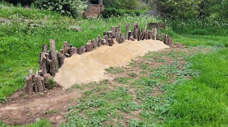 A series of upright logs, buried into the ground with sand/earth built up against them to create a bank for bees to nest in