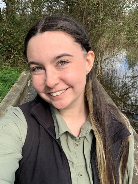 A photo of staff member Amy. She has long dark hair, is smiling and wearing a green shirt and gilet