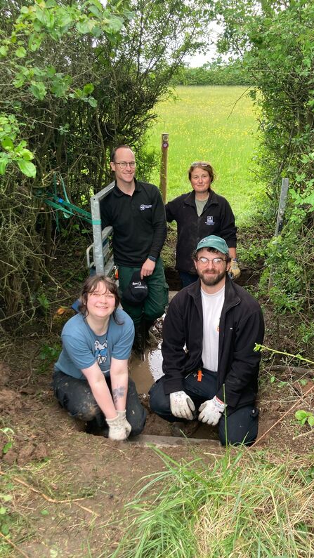 A photo of four conservation trainees crouching in front of a small bridge over a stream with hedgerow behind smiling at the camera
