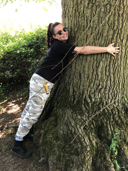 Amy is hugging the trunk of a large tree - she's looking at the camera and has a big smile on her face