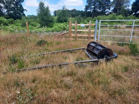 A photo of a large roller laid down in a field with fence and bracken behind