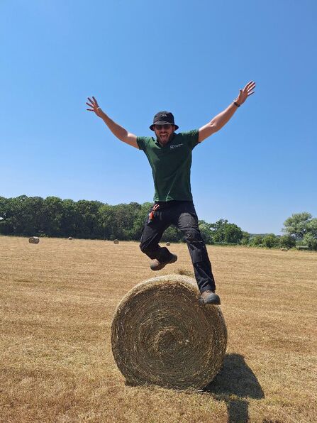 Person leaping off a round hay bale in a sunlit dry field, arms outstretched and smiling, wearing a bucket hat, green T‑shirt and black trousers with other hay bales and tree line in the distance.
