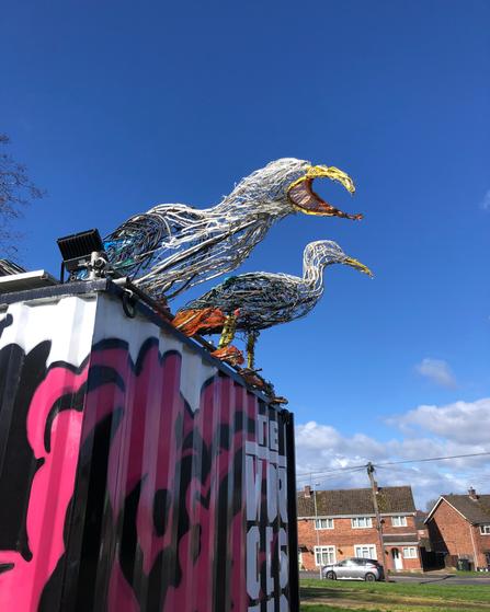 A photo of sculptures of two gulls made of old wire standing on top of a large black plinth box with blue sky in the background
